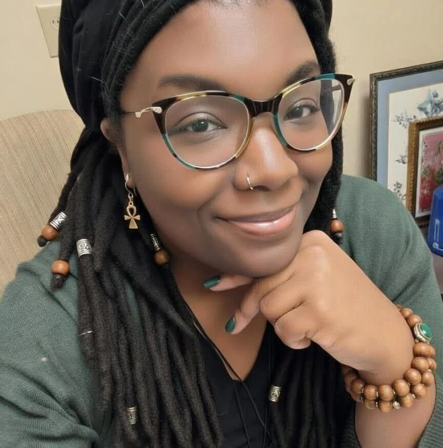 A smiling woman with dark skin, glasses, and dreadlocks wearing a gray shirt, yellow cardigan, and a silver necklace, standing against a plain light-colored background.