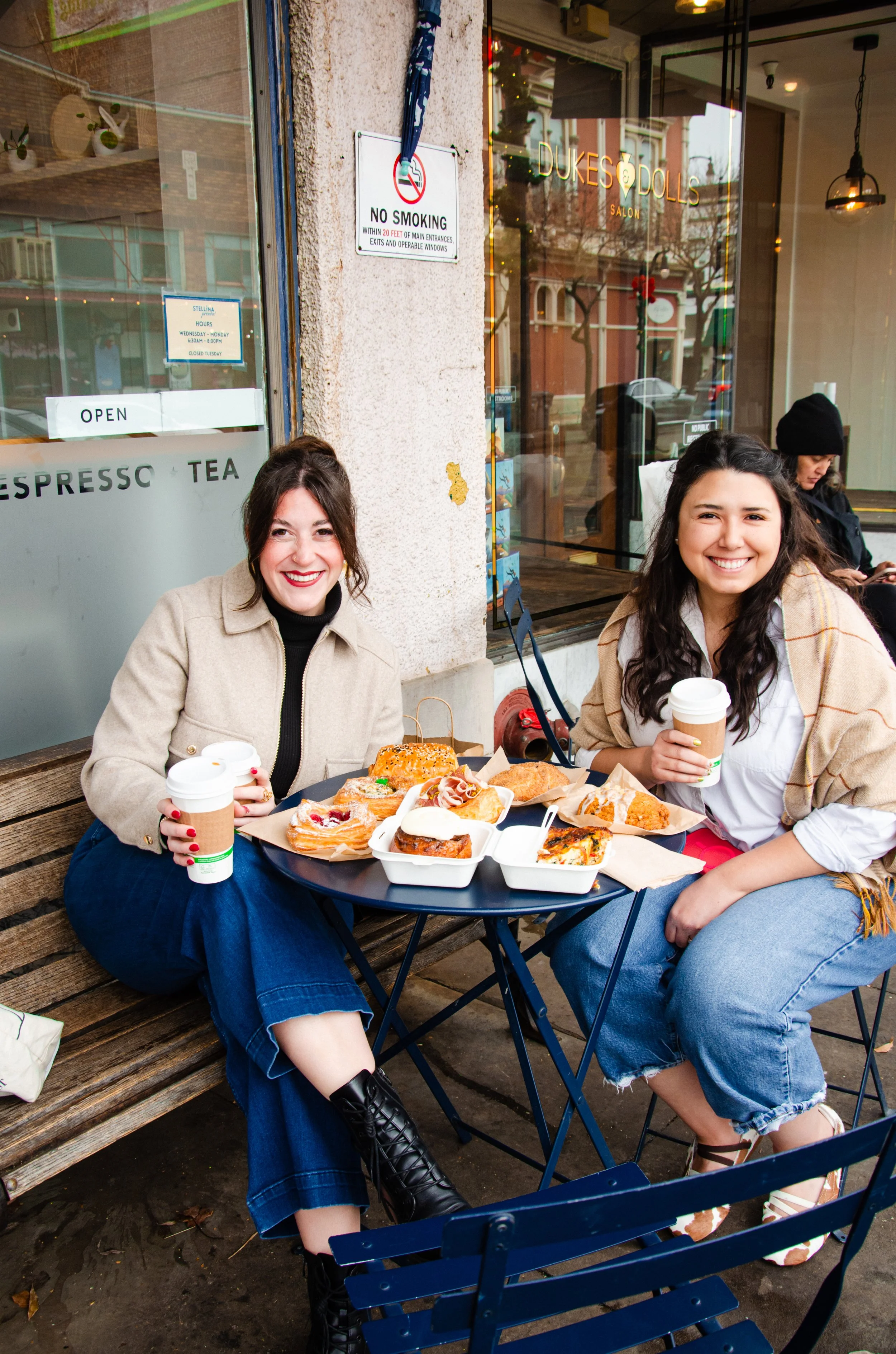 Emma's sister Camille who has brown hair and a red lipstick smile is sitting with her friend Victoria who has long brown hair and is smiling as well. They are holding coffee cups and smiling as they sit at a table full of pastries.