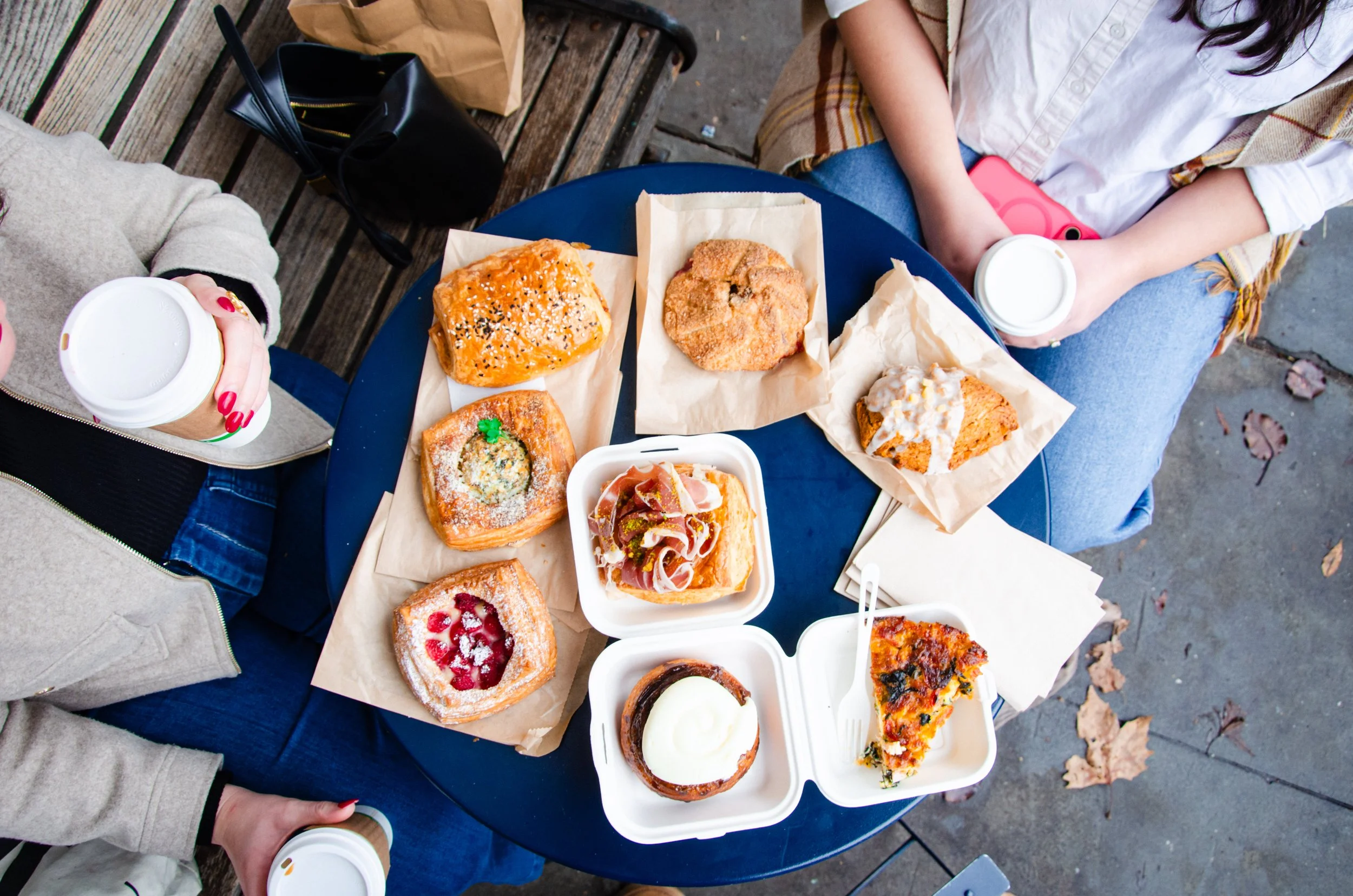 an overhead shot of the table with many pastries. Two people are partially in the frame, each holding coffee cups.