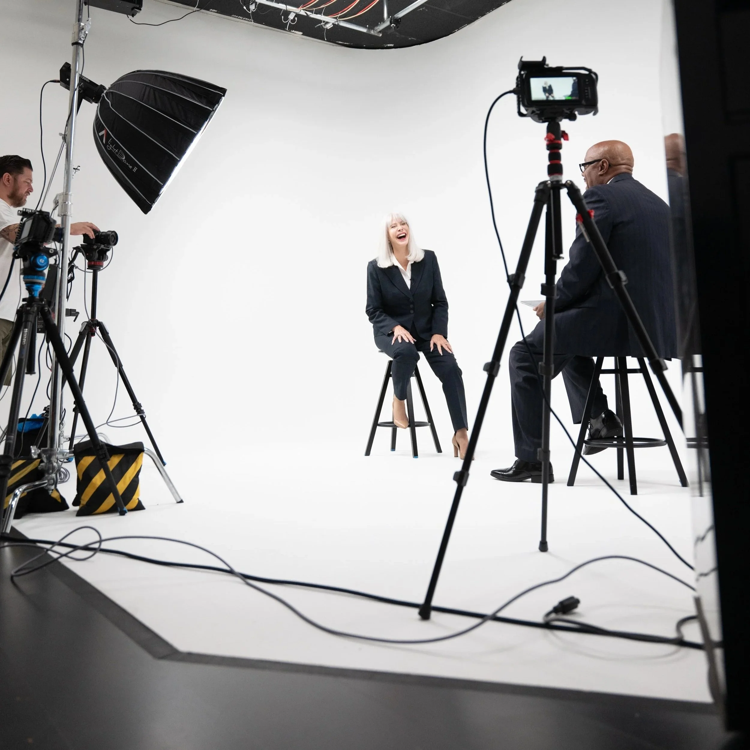 A woman in a black suit is sitting on a stool, laughing, during a video shoot in a white studio. Two men, one with a camera and the other with a microphone, are recording her, along with various lighting and filming equipment around them.