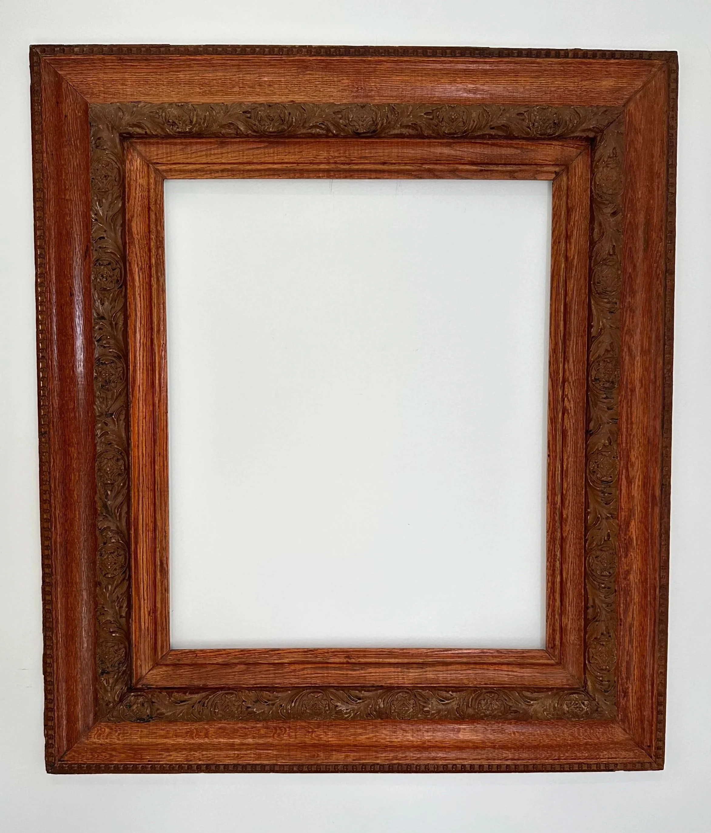 Empty wooden picture frame with ornate carvings hanging on a white wall.