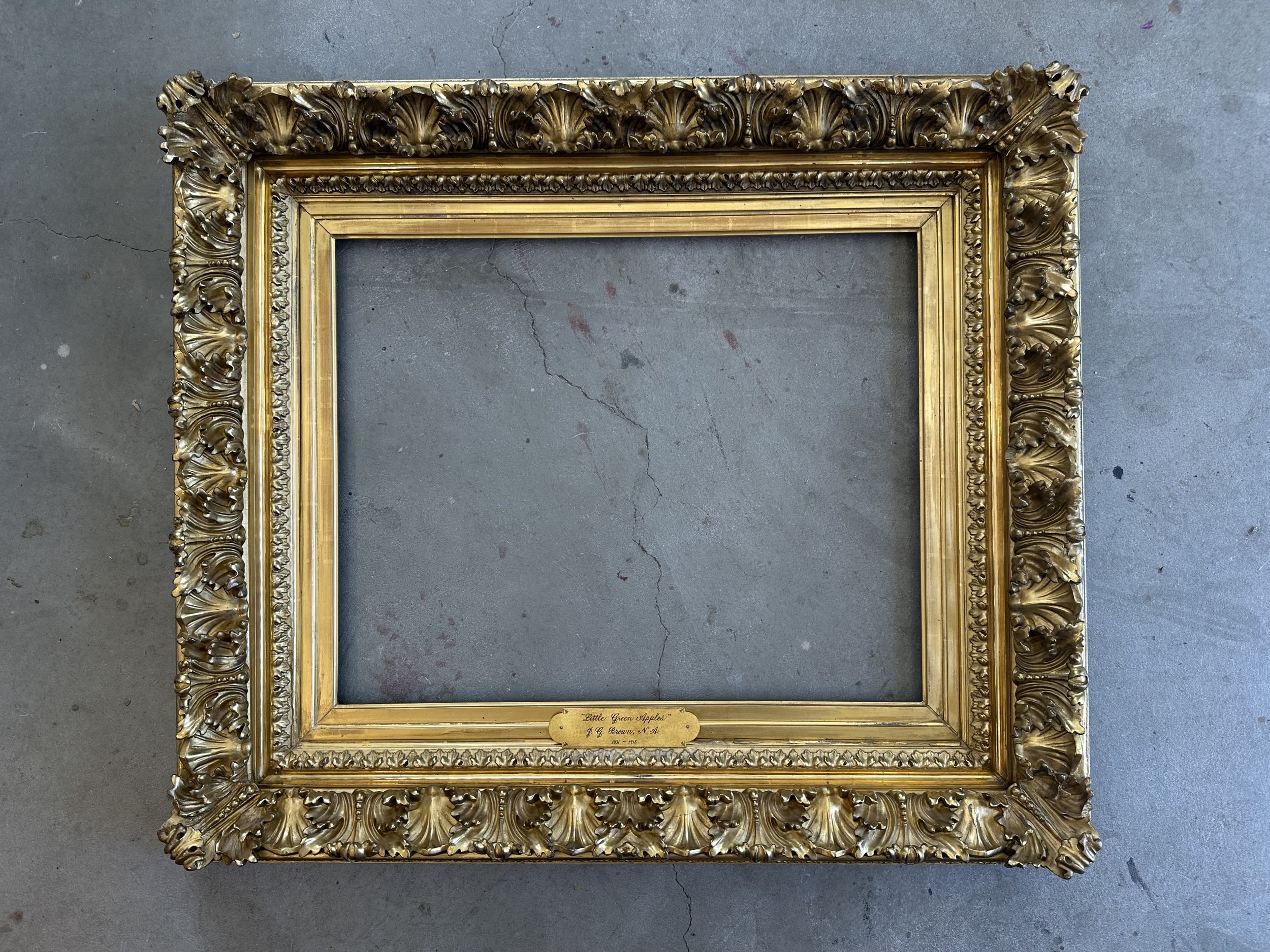 An ornate empty gold-colored picture frame with intricate details on a concrete floor. A small plaque on the bottom of the frame reads 'Little Green Apple J. H. Brown, N. J.'
