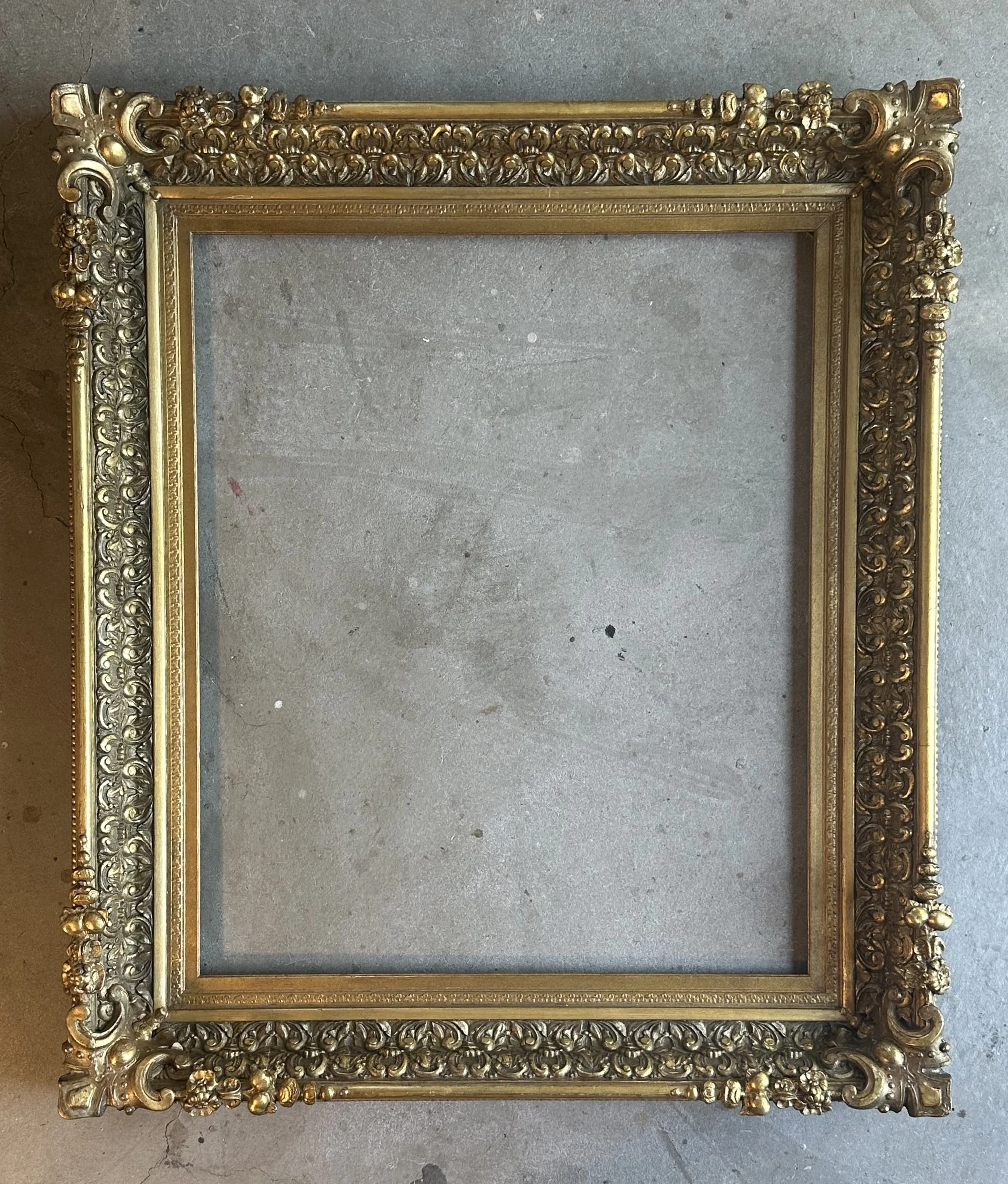 An ornate gold picture frame with intricate floral and scroll designs, lying on a concrete floor.