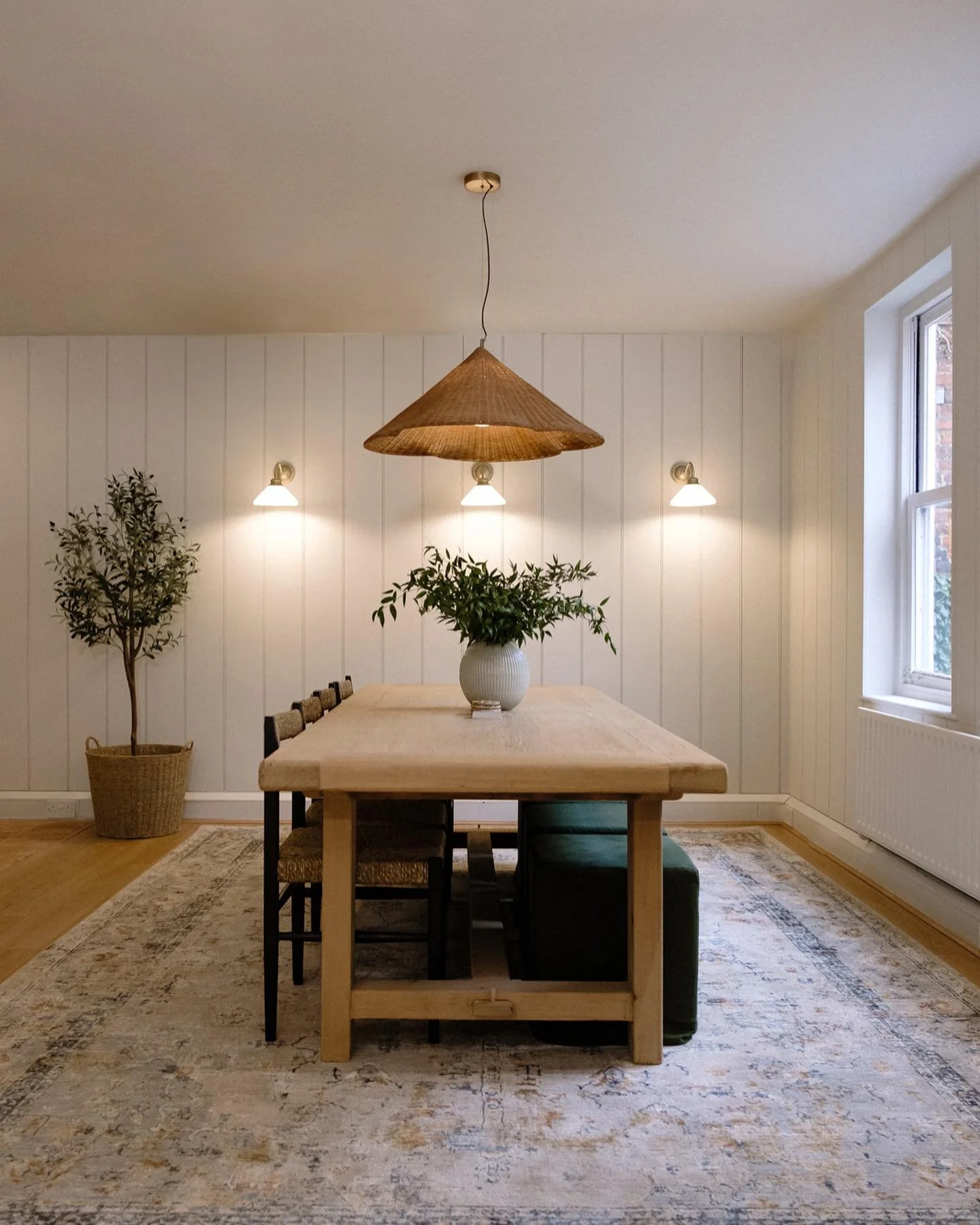 Dining room with wooden table, wicker pendant light, and wall sconces. Chairs are arranged around the table, a potted plant on top, and a large basket with a plant beside it. The room has a light-colored rug and paneled walls.