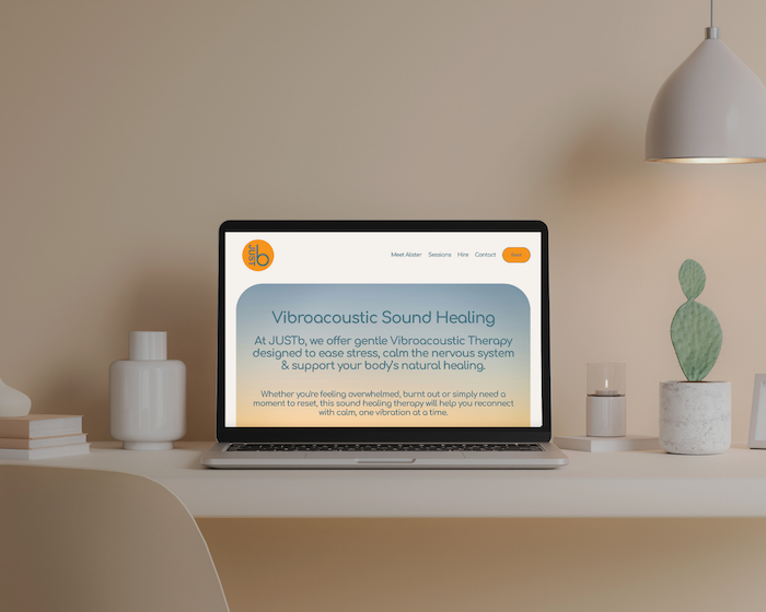Laptop on a white desk displaying a website about Vibroacoustic Sound Healing, with minimalist decor including a white vase, a stack of books, a cactus in a pot, and a white lamp.