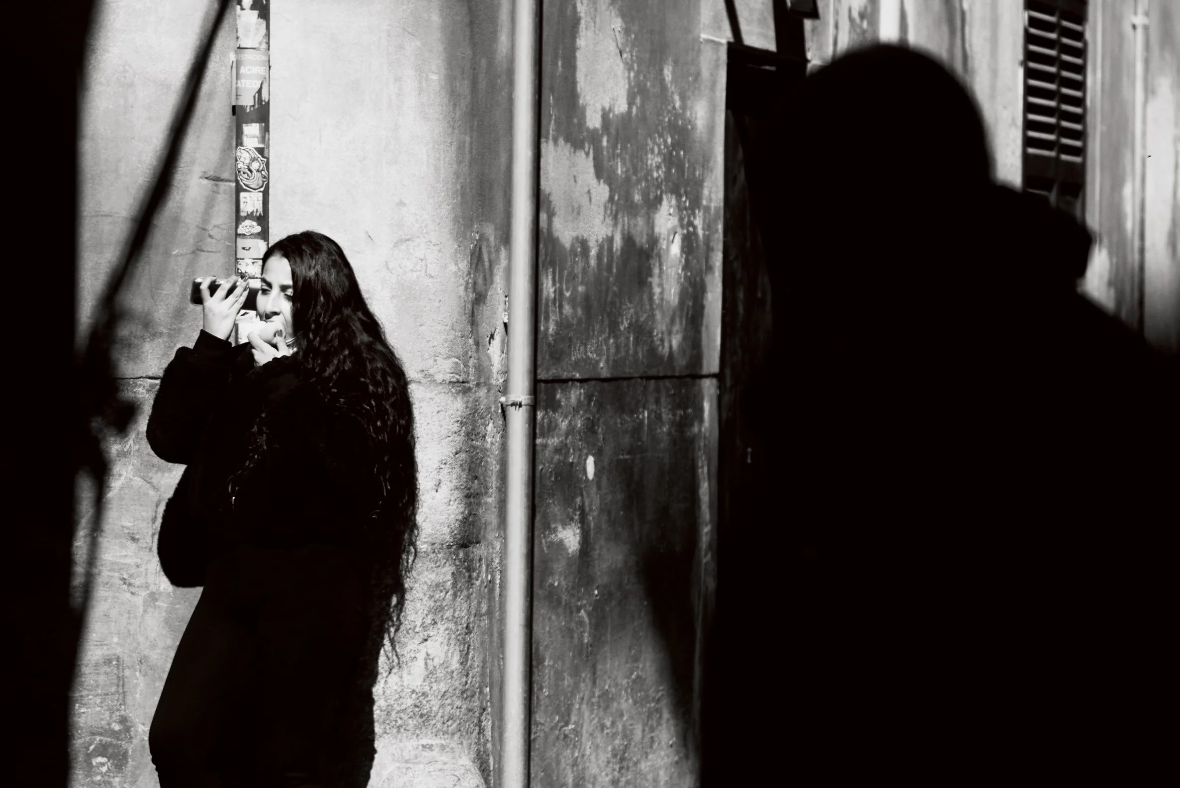 A woman with long dark curly hair taking a photo with her phone against a rough concrete wall, while a large shadow of a person is cast on the wall nearby.