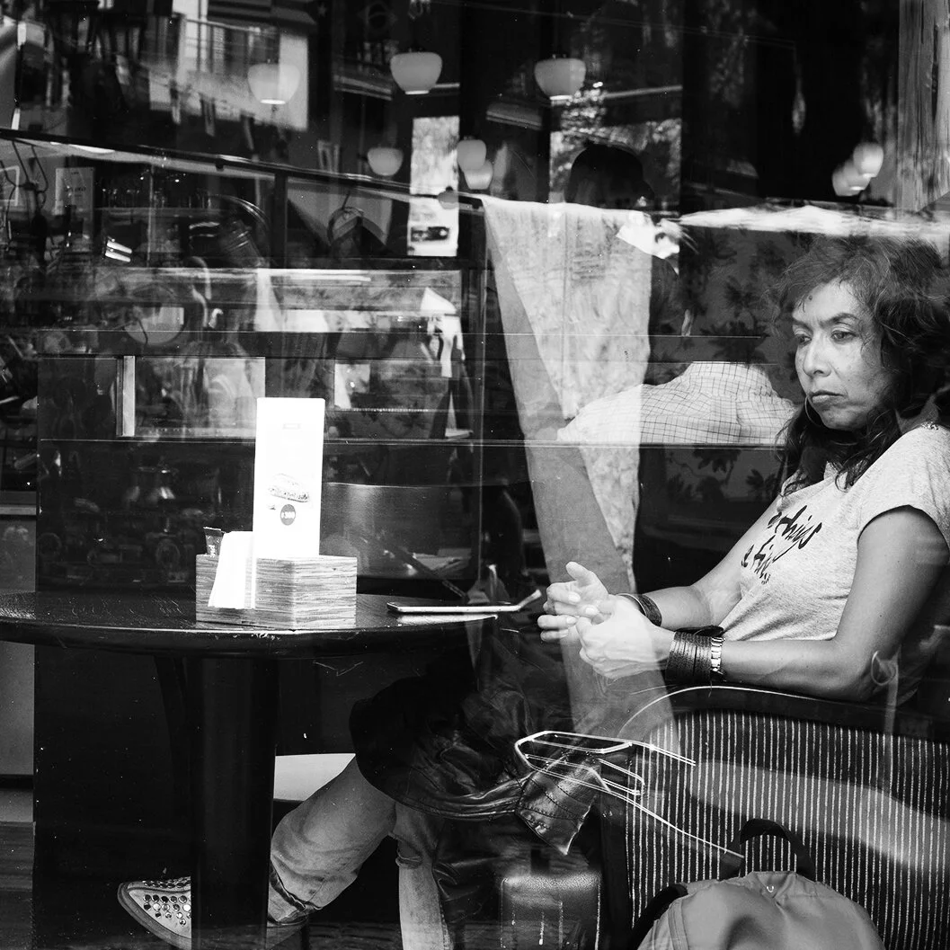 A woman sitting alone inside a cafe, viewed through a glass window with reflections of the street outside. She looks contemplative, with her hands clasped, and has a bag and a phone on the table in front of her.