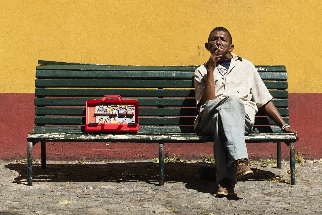 A man sitting alone on a green park bench, smoking, with a red case of snacks beside him, in front of a yellow and red wall.