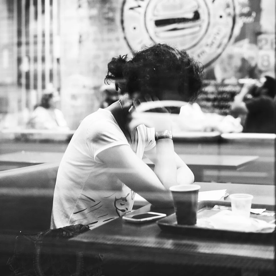 A person with curly hair sitting at a table in a coffee shop, leaning on their arm with their head resting on their hand; there are cups, a smartphone, and a tray on the table, and other people are visible in the background through the window.