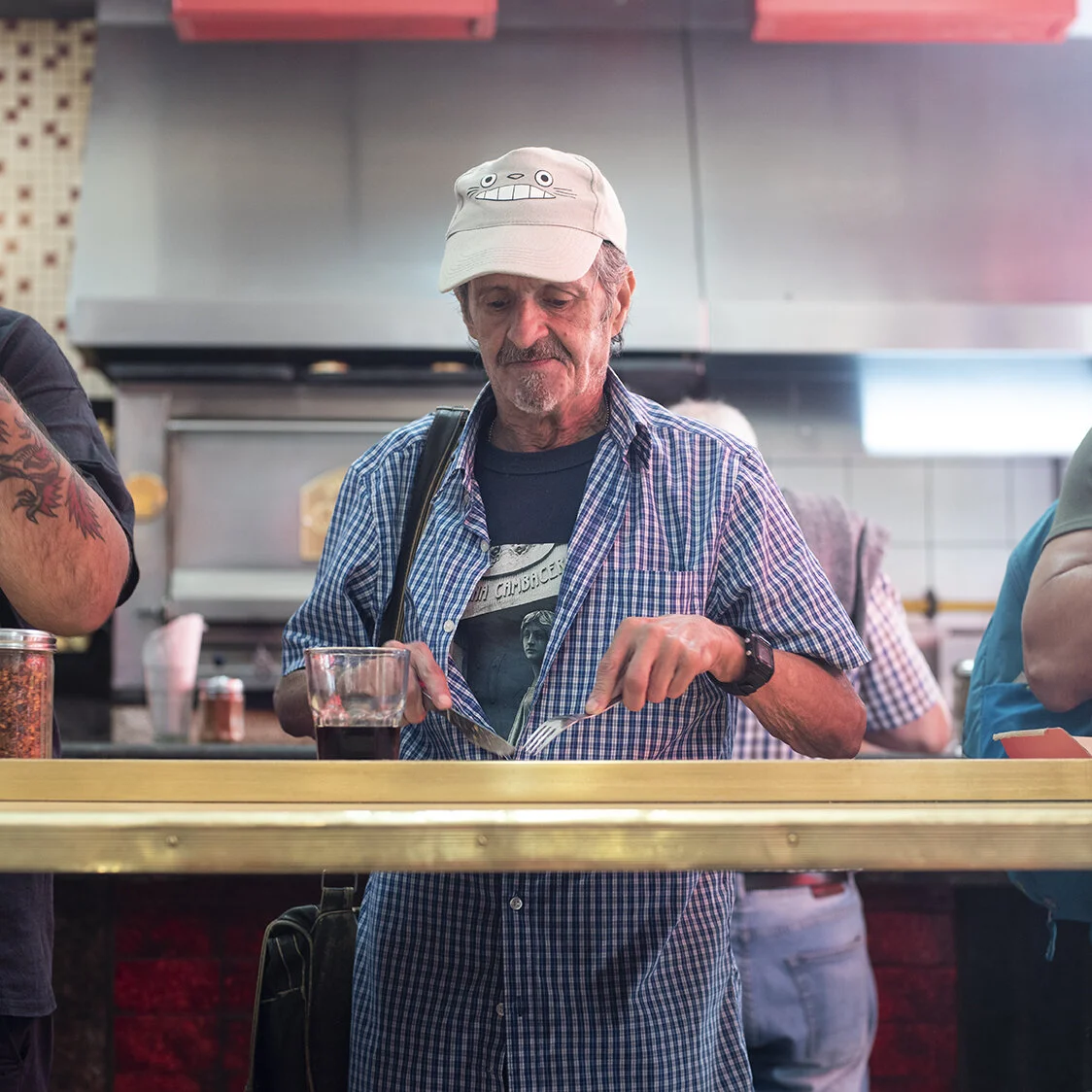 An elderly man with a plaid shirt and a cap with a cartoon face on it is eating with a fork at a food counter. There is a glass of dark soda in front of him, and he is in a casual dining setting.
