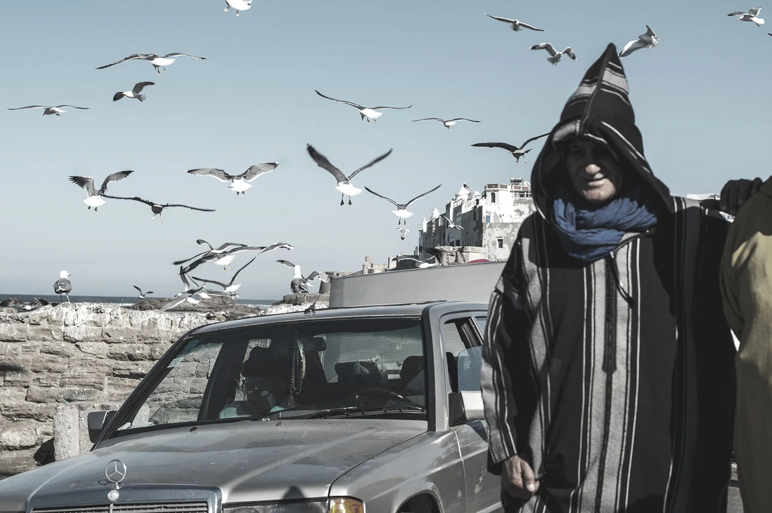 A person in a hooded striped jacket and blue scarf walking past a silver Mercedes-Benz car, with seagulls flying in the background near a stone wall and white buildings.