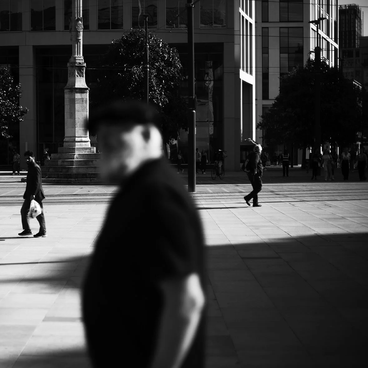 A blurred person in the foreground with a city street and several pedestrians in the background, featuring modern buildings and a statue in black and white.