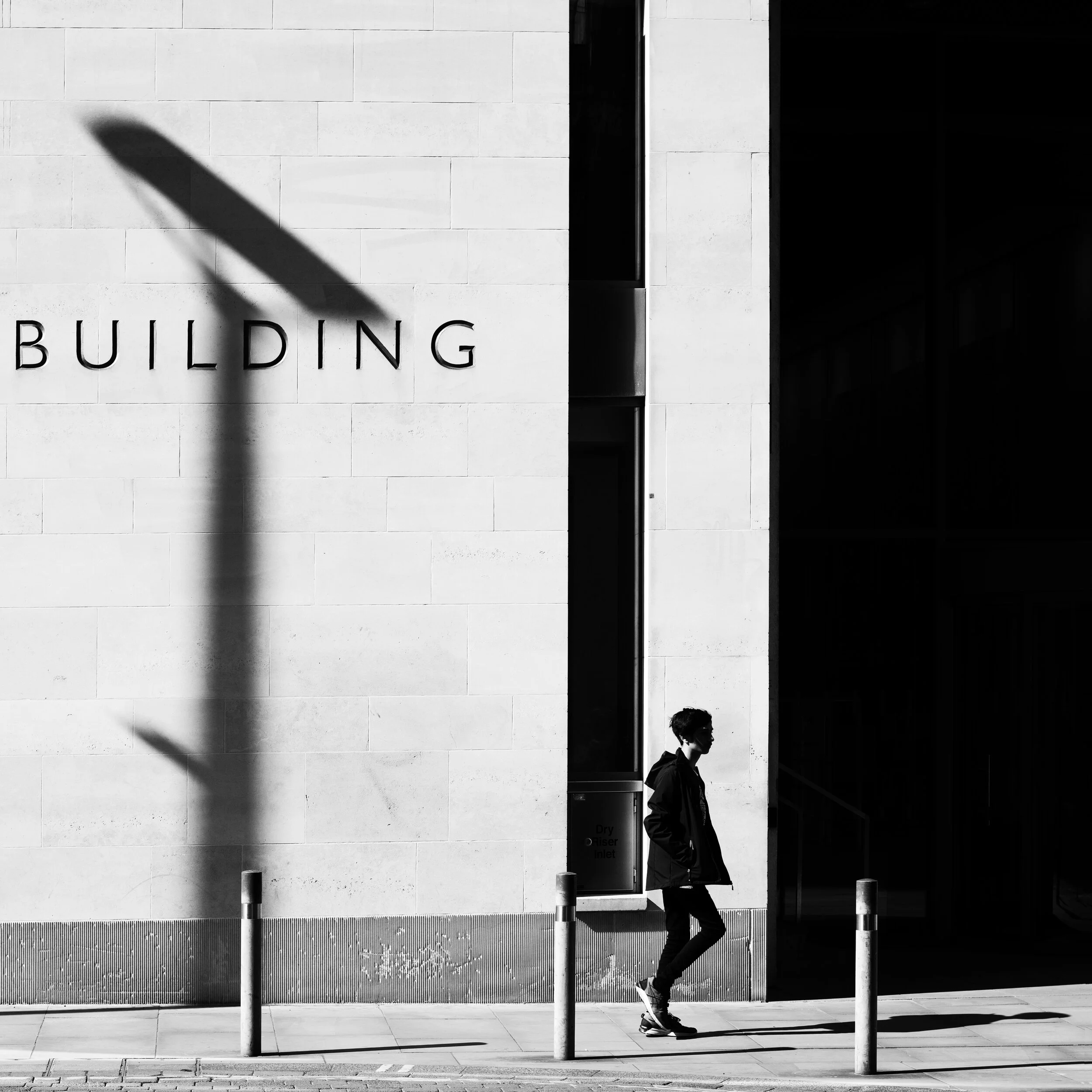 A person walking along a sidewalk in front of a building with a shadow of a streetlamp cast on the wall. The building has the word 'BUILDING' on its exterior.