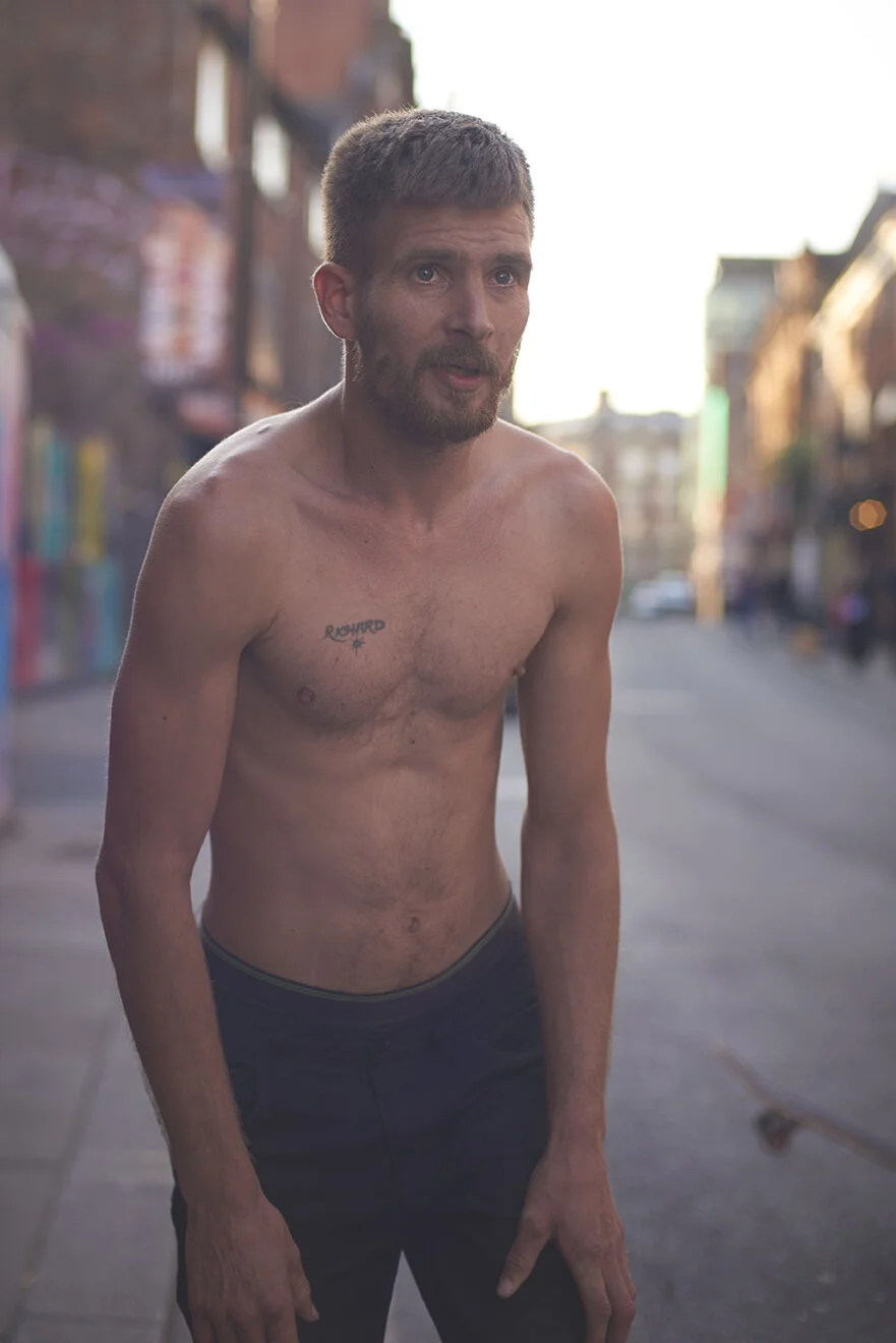 A shirtless skateboarder with a beard and short hair standing on an urban street during daylight, with buildings and trees in the background.
