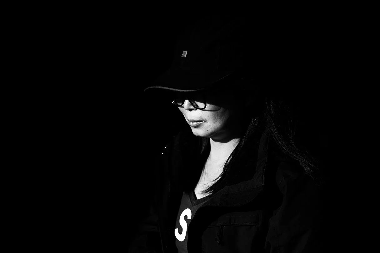 A woman wearing glasses and a hat in a dark setting, with her head slightly bowed.