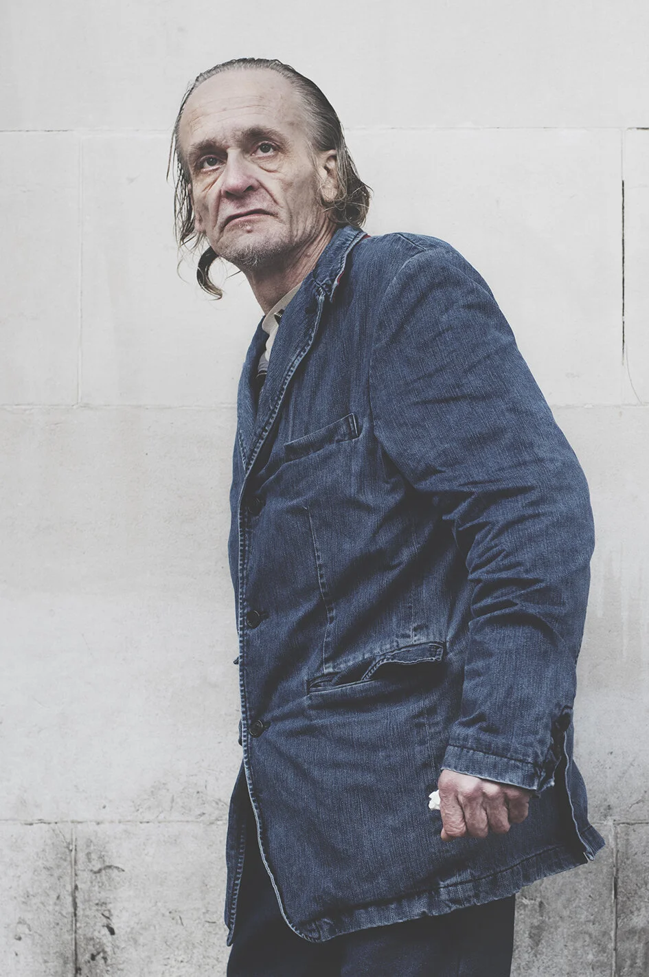 An older man with long, gray hair, wearing a blue denim jacket, standing against a plain white wall with a serious or contemplative expression.