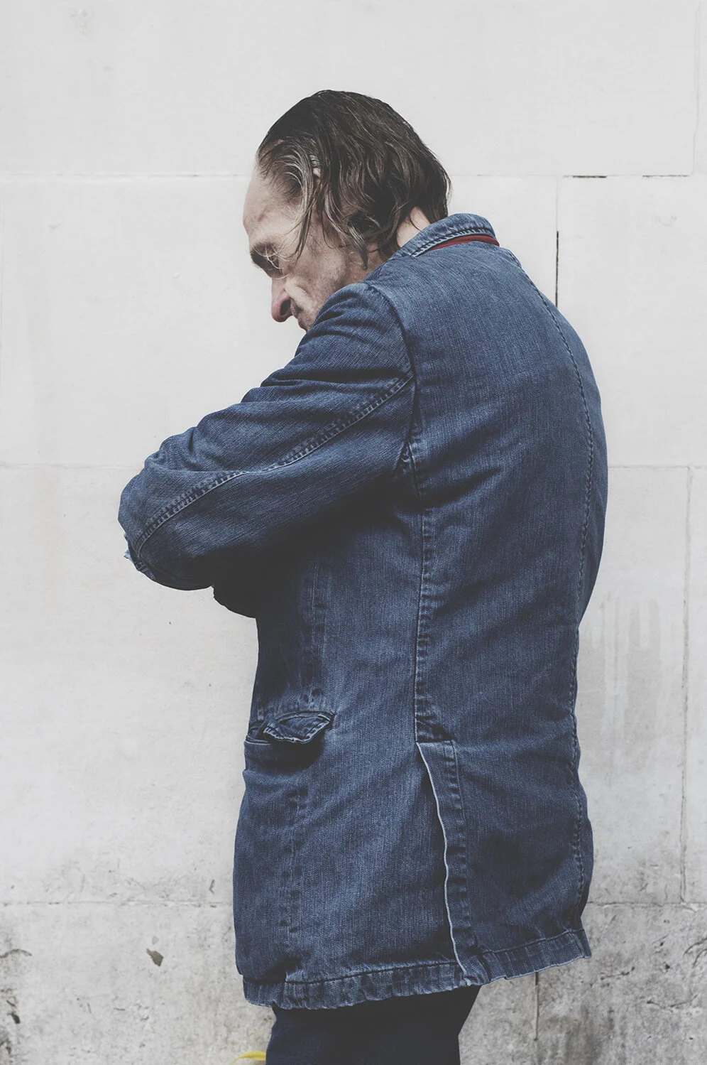 A man with long hair wearing a denim jacket standing against a plain white wall, looking down with his arms crossed.