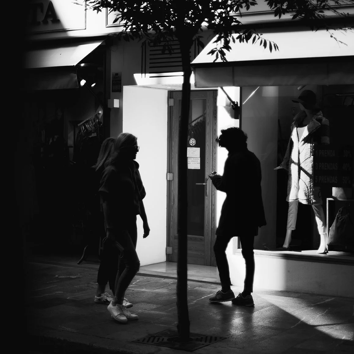 Three people standing and talking outside a store at daytime, with another person visible inside through the display window. The scene is in black and white, with shadows cast natural lighting.