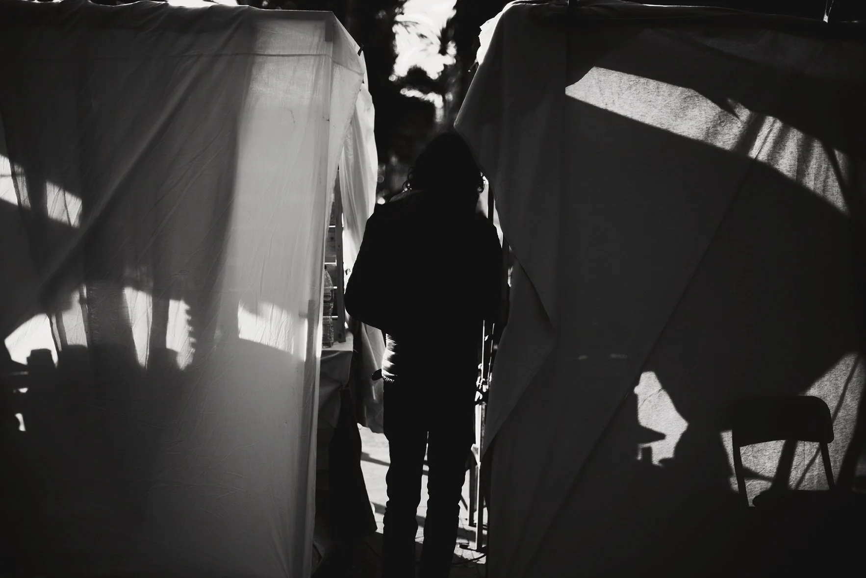 A person silhouetted against sunlight standing between two fabric tents or booths, with shadows cast on the tents and a chair inside one of them.
