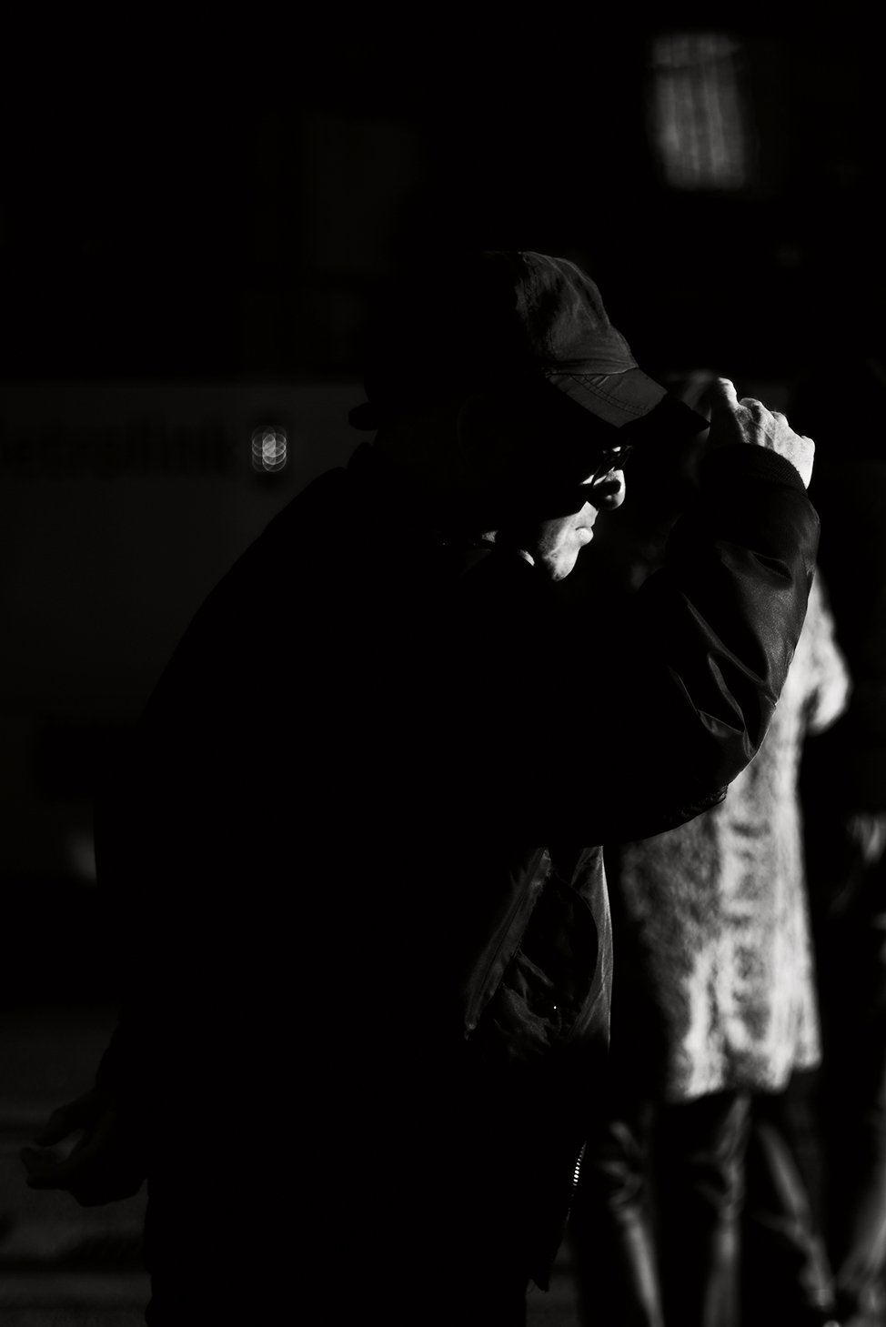 A silhouette of a man wearing sunglasses and a cap, adjusting his cap in silhouette against a dark background.