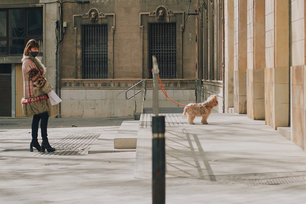 A woman wearing a face mask and a patterned coat stands on a city sidewalk queueing up at a bank behind a dog, holding a paper while looking at the small dog on a leash. The dog is standing near a building with stone walls and metal railings.
