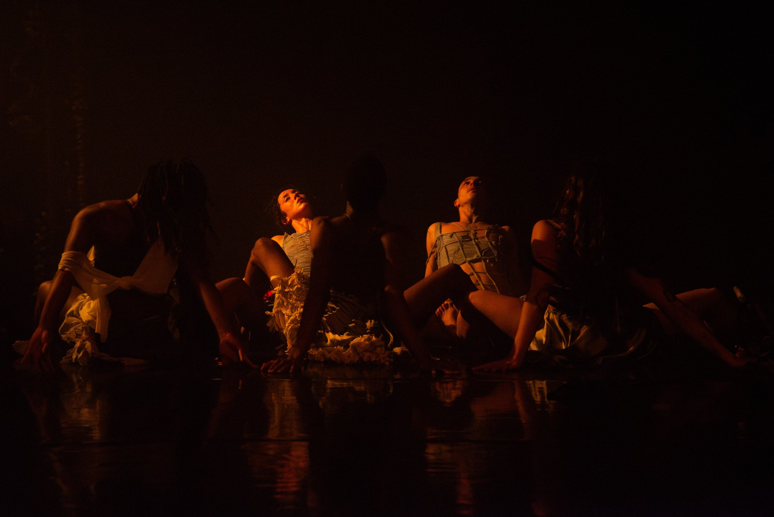 A group of dancers sit in a circle with their legs outstretched in a shadowy space.