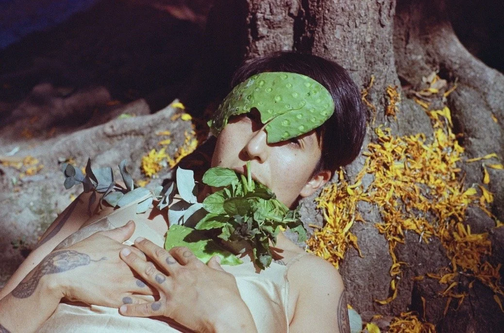 Joan Gutiérrez reclines against a tree adorned with various plants, a cactus framing her head, herbs in her mouth, and leaves around her shoulders and collarbones. Her tattooed hands are clasped at her chest.