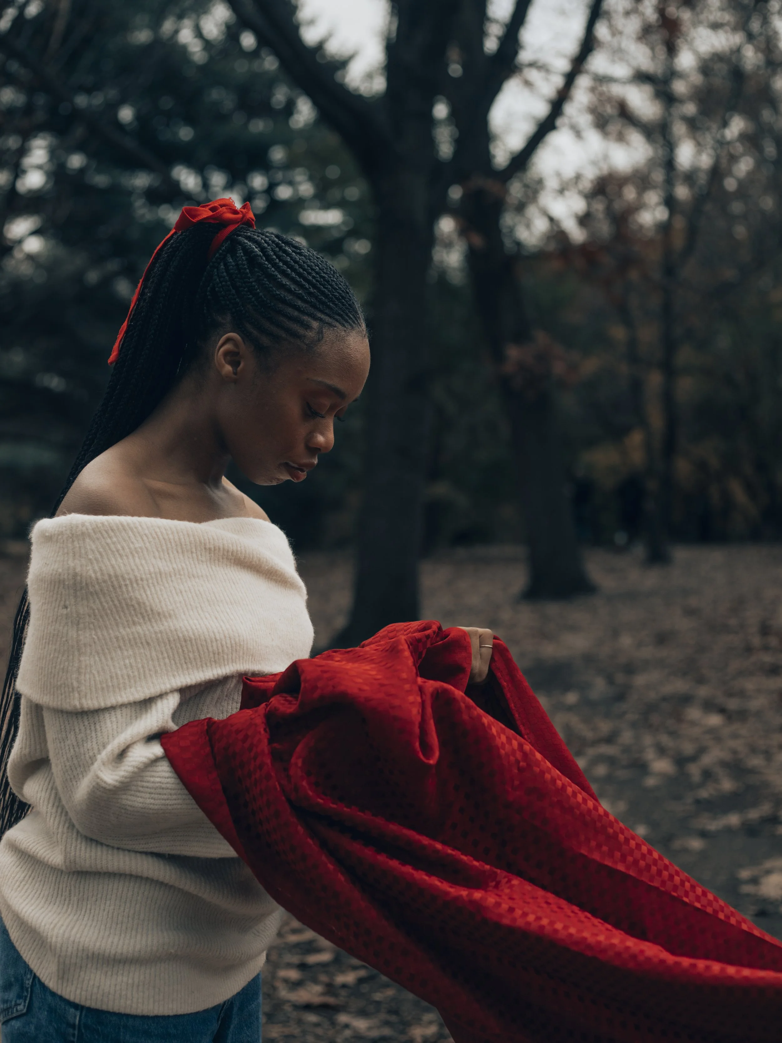 A young Black woman with braided hair and a red ribbon holds a flowing red fabric outdoors in a wooded park.