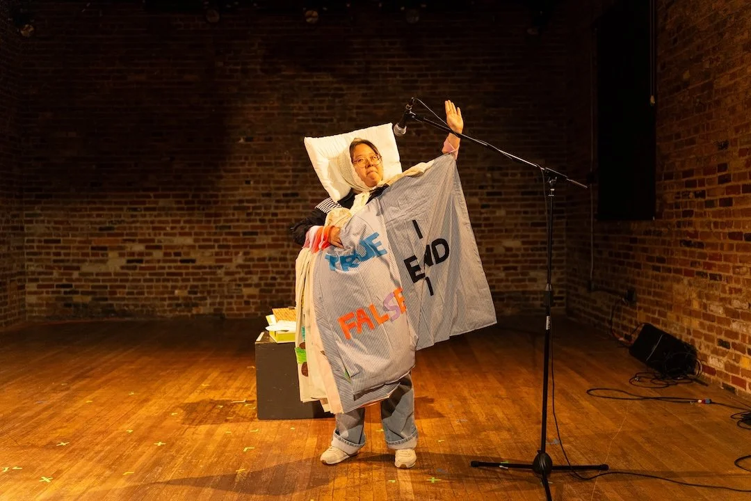 Yiseul LeMieux stands on a hardwood floor against a red brick backdrop, raising her arm while dressed in a costume seemingly made out of pillows and bedsheets. The words "True" and "False" are visible on the costume.