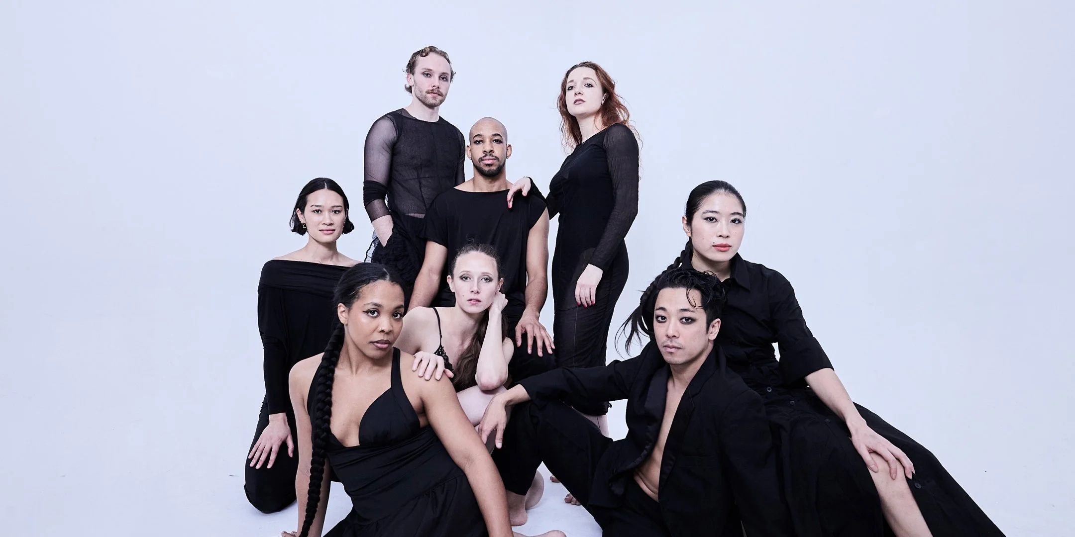 A studio portrait of nine adults posed together against a seamless white background. They wear coordinated black clothing in varied textures—sheer mesh, sleeveless tops, fitted dresses, and layered separates—creating a unified yet individual look.