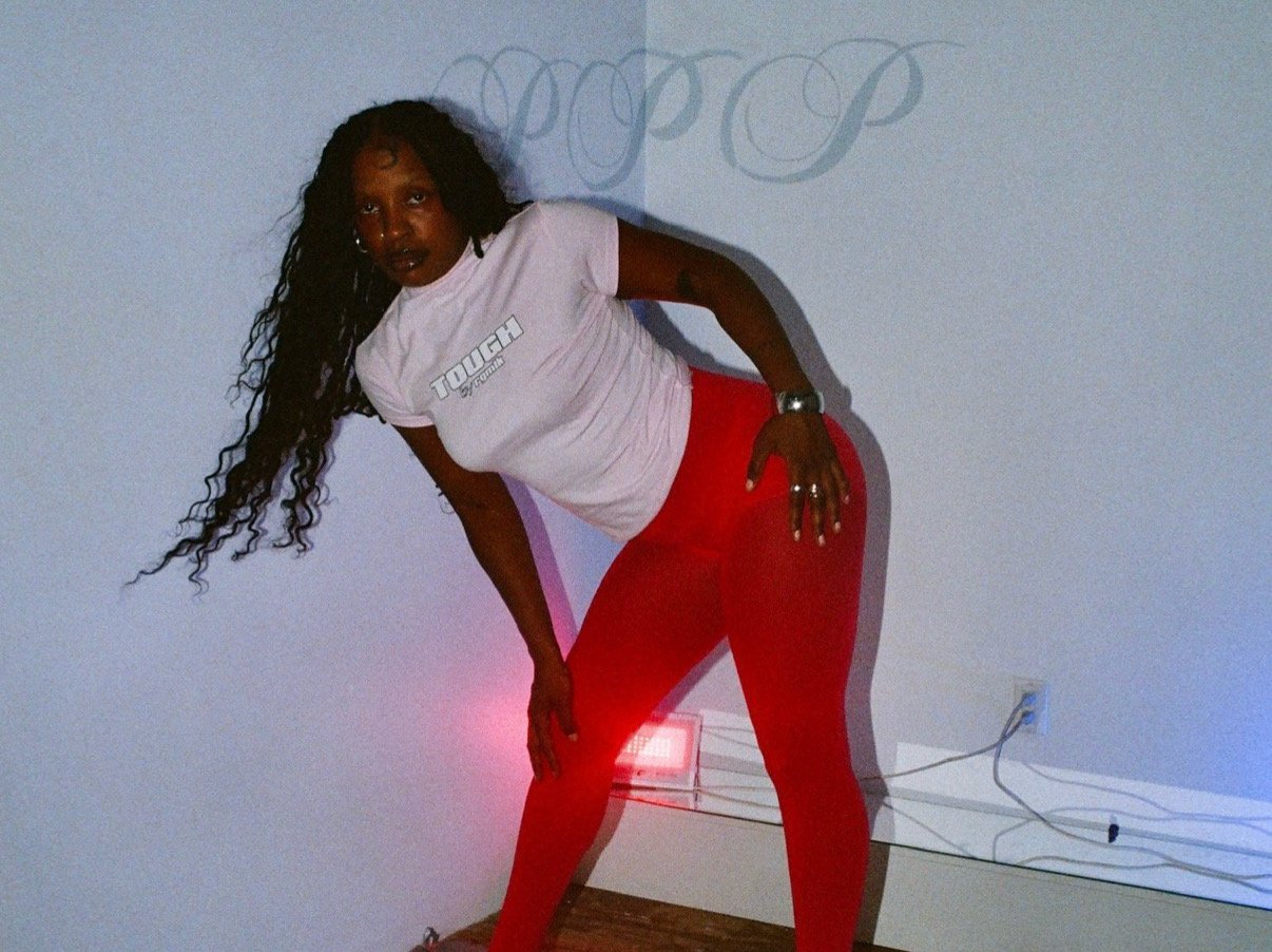 Joy Norton leans to the side in red tights and white shirt printed with the word "TOUGH"