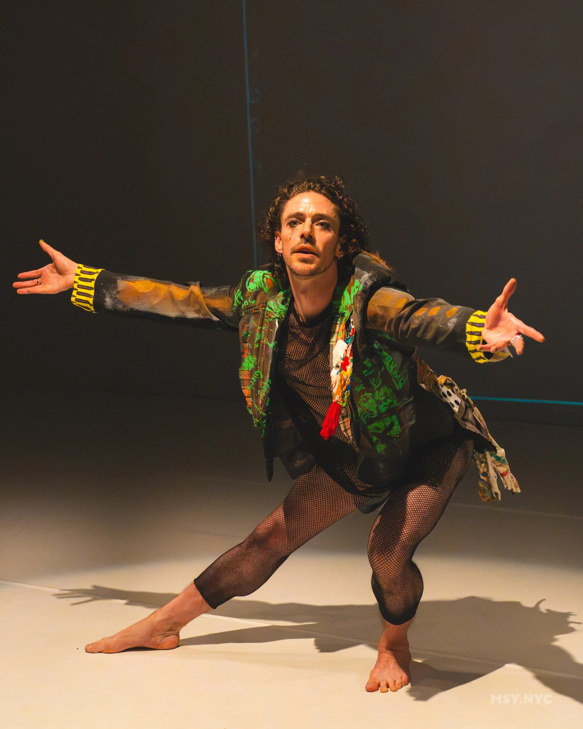A dancer in a painted black jacket painted with yellow and green wearing a mesh top and fishnet stockings, gestures towards the camera in a bow formation, arms outstretched.