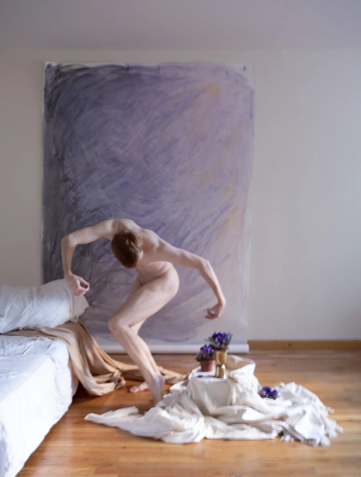 Nude male dancer with short ginger hair doubled over with beveled legs and arms curved out to the sides. The dancer is in an apartment with a couch with a white sheet on it, a purple pastel colored wall hanging, and potted violets on a white sheet.