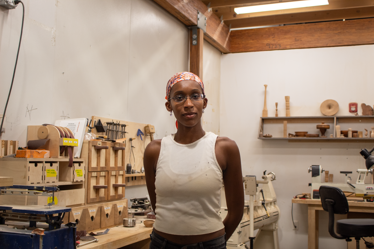 Nifemi Ogunro, wearing a white tank top, glasses, and a red patterned headwrap, stands before the camera in a studio space full of various wooden objects.