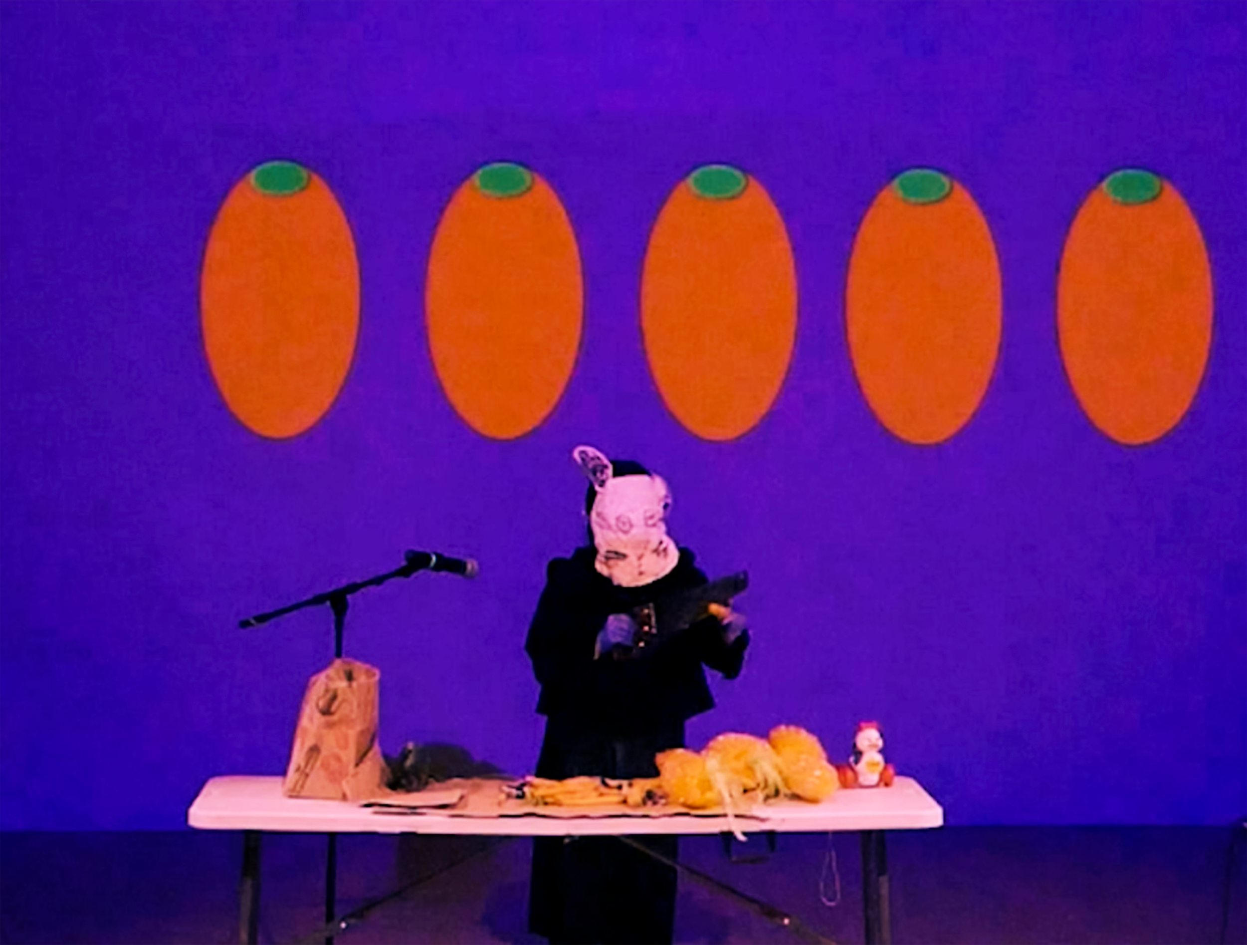 A figure in a handmade rabbit mask is using a handsaw to cut through a small object. The figure stands behind a table full of plastic toy figures and a grocery bag, against a purple backdrop with 5 abstract oblong renderings of carrots in a row.