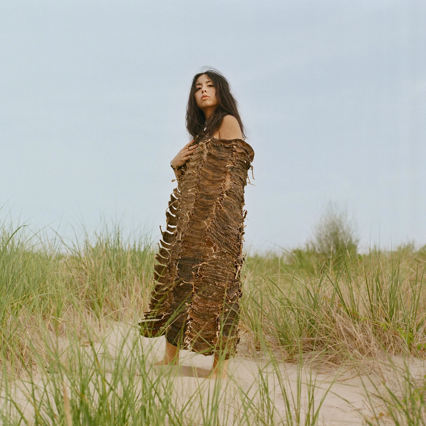 Erica Enriquez stands facing the camera in a dress made of multiple banana peels stitched together with white thread, against a sandy dune backdrop with tall green grass and a cloudless pale blue sky.