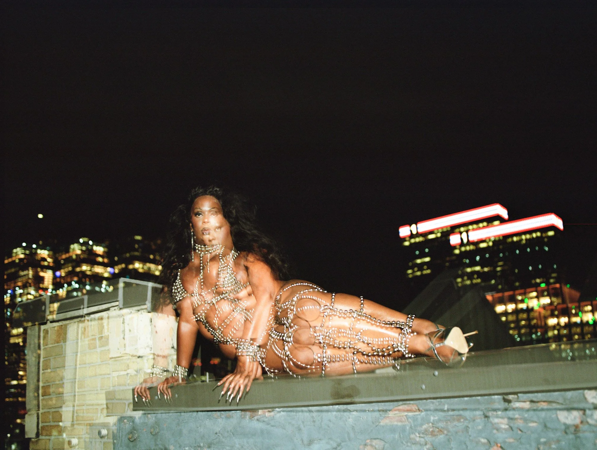 DANIRO, in a double exposure photograph, reclines at the edge of a tall building against a nocturnal city backdrop. Daniro is wearing a pearl harness bodysuit and heels, and her white acrylic nails are gripping the edge of the building.