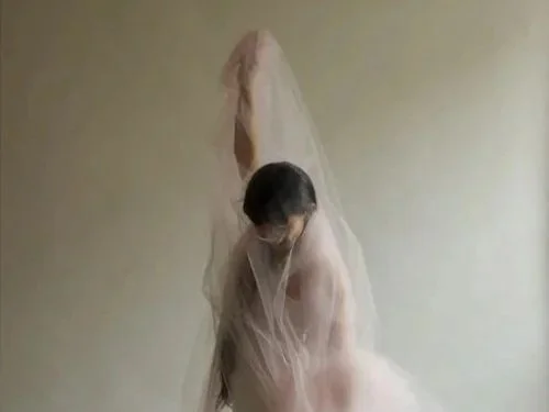 Yuki Kawahisa stands with one arm raised above her head, covered in white translucent fabric, against a muted grey backdrop.
