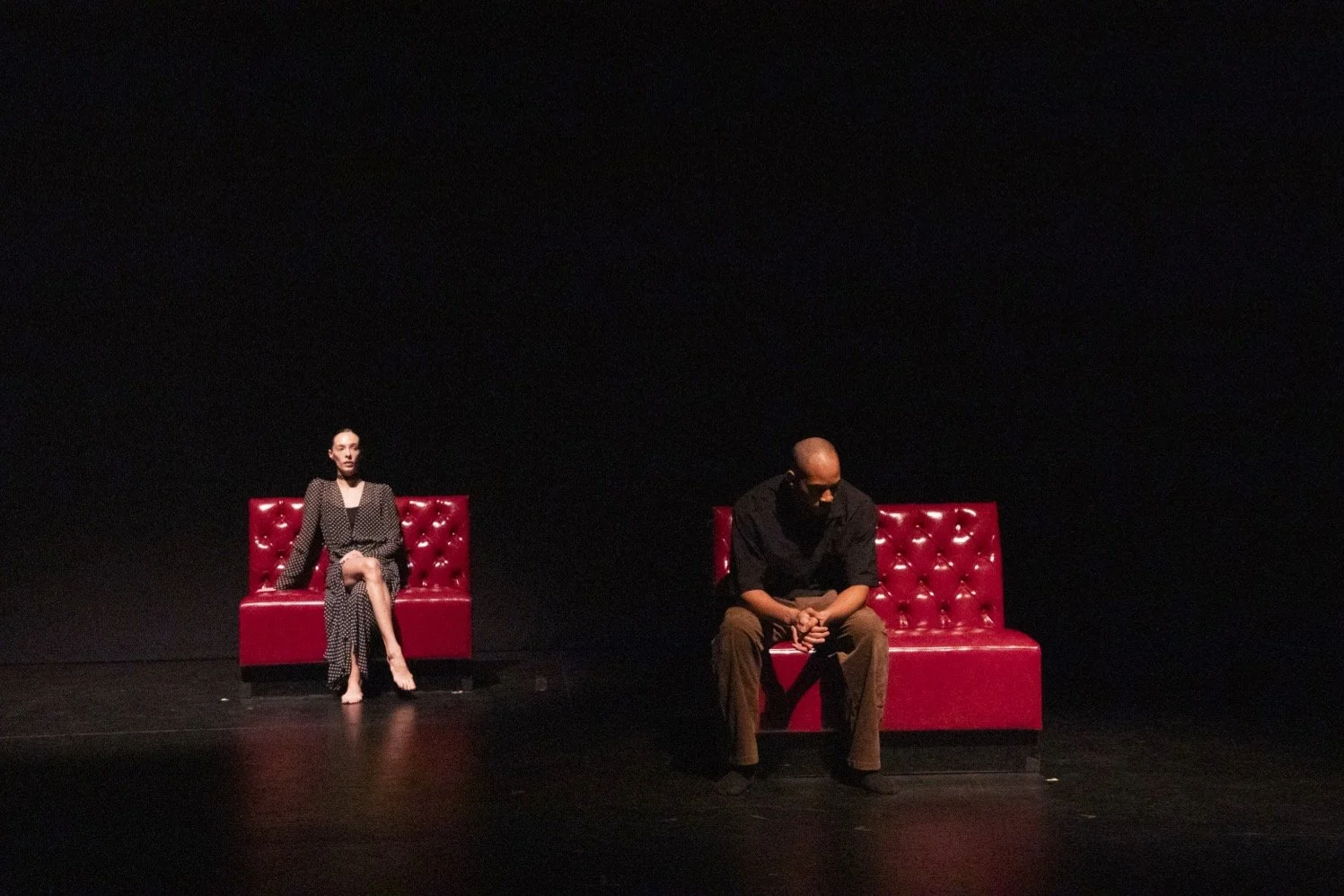 Two performers, one downstage and one upstage, sit on shiny red vinyl couches.