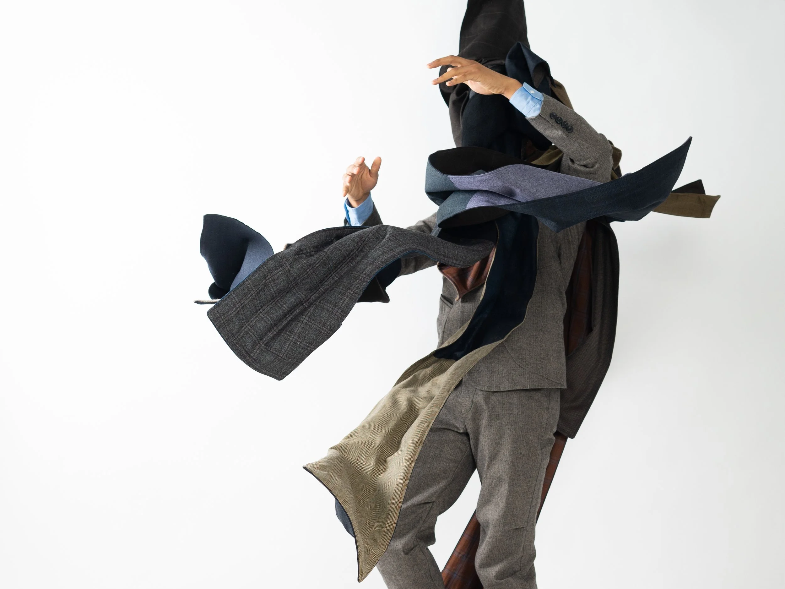 Quaba Venza Ernest raises his arms mid-motion against a white background. He is wearing multiple scarves and jackets that obscure his face.