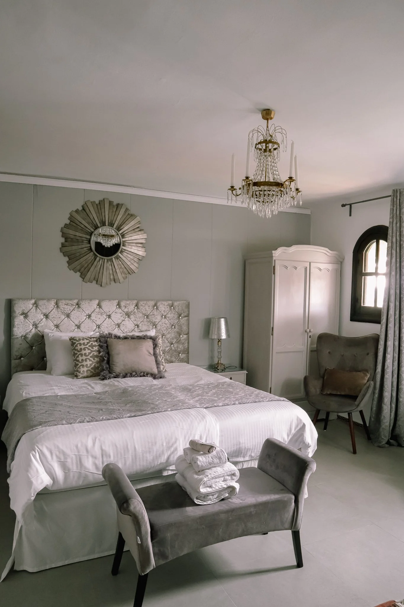Elegant bedroom with a large bed featuring a tufted silver headboard, decorative pillows, and a folded towel set at the foot. There is a chandelier overhead, a bedside table with a silver lamp, a white wardrobe, and an armchair near a window with curtains.