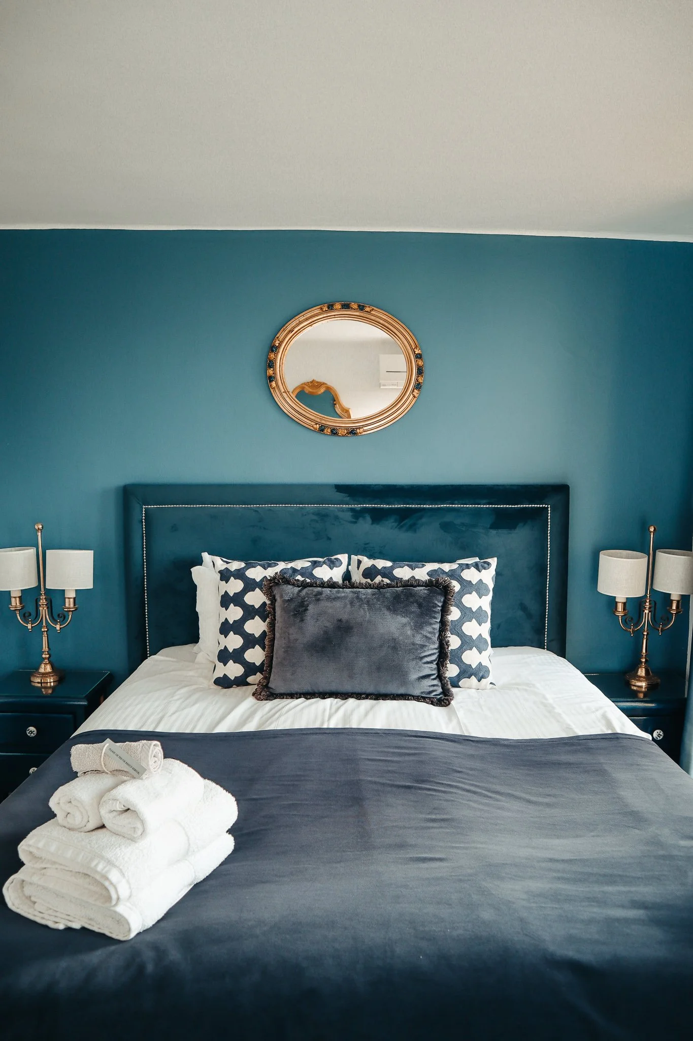 A neatly made bed with dark blue bedding, decorative pillows, and a stack of white towels on the bed, in a bedroom with blue walls, gold-accented lamps, and a round mirror above the headboard.