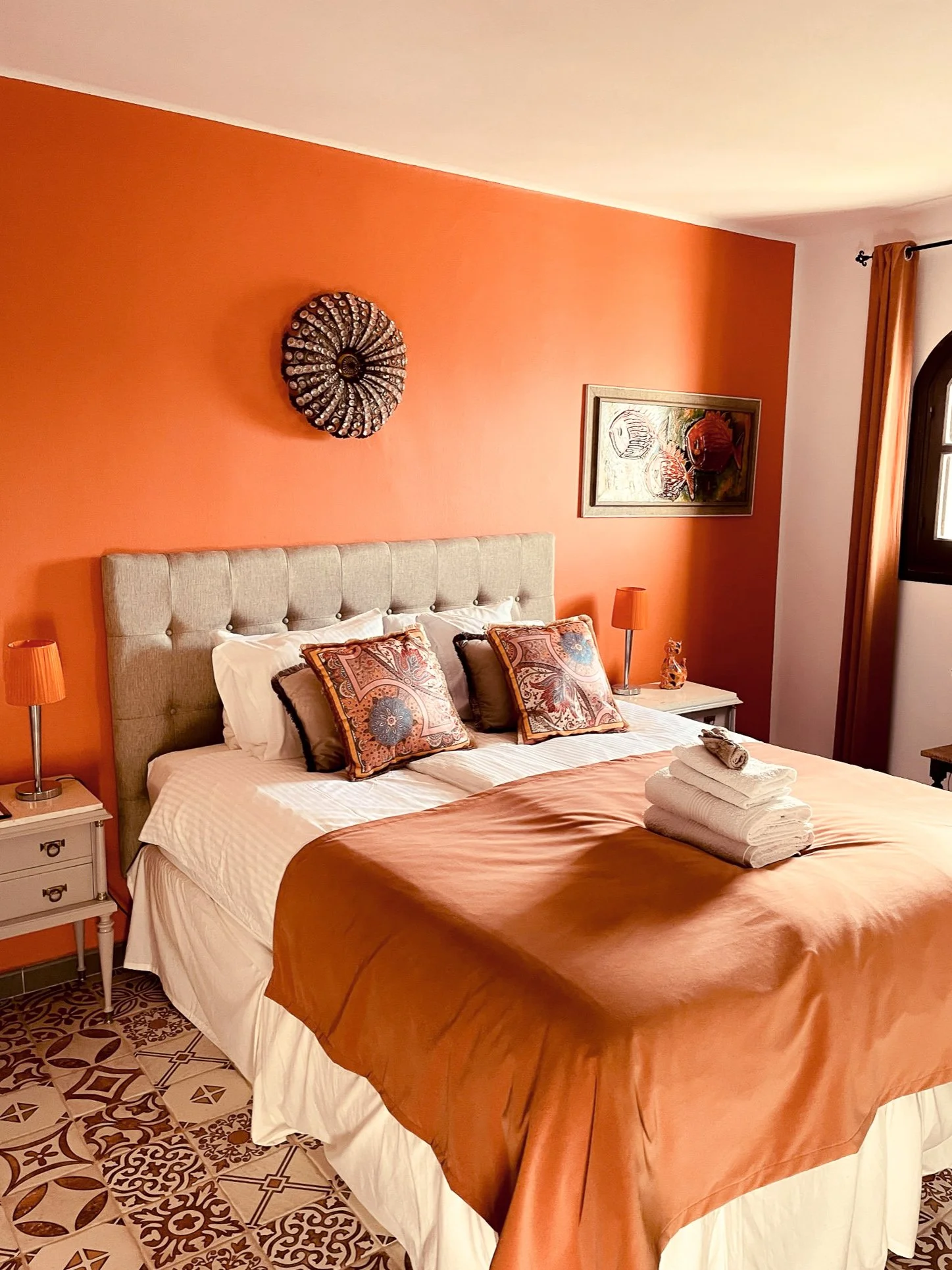 A neatly made bed with a beige tufted headboard, orange and patterned decorative pillows, and an orange bedspread with a stack of white towels on top. The room has orange walls with wall art and a circular decorative piece, white nightstands with orange lamps, and patterned floor tiles.