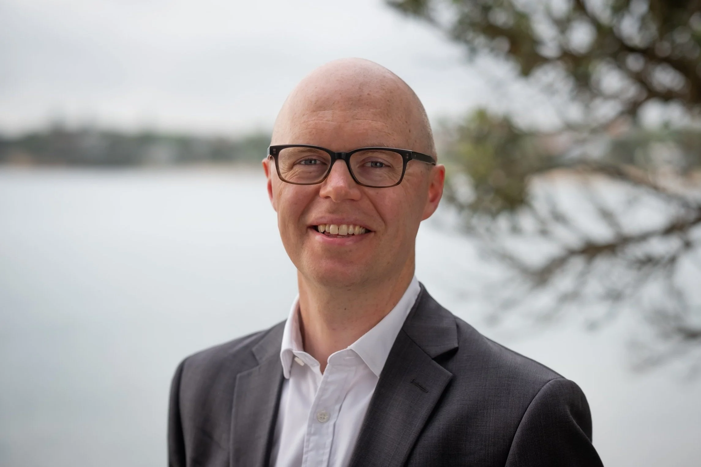 Colin Downie - A man with a shaved head, glasses, wearing a dark blazer and white shirt, smiling outdoors near water with blurred trees in the background.