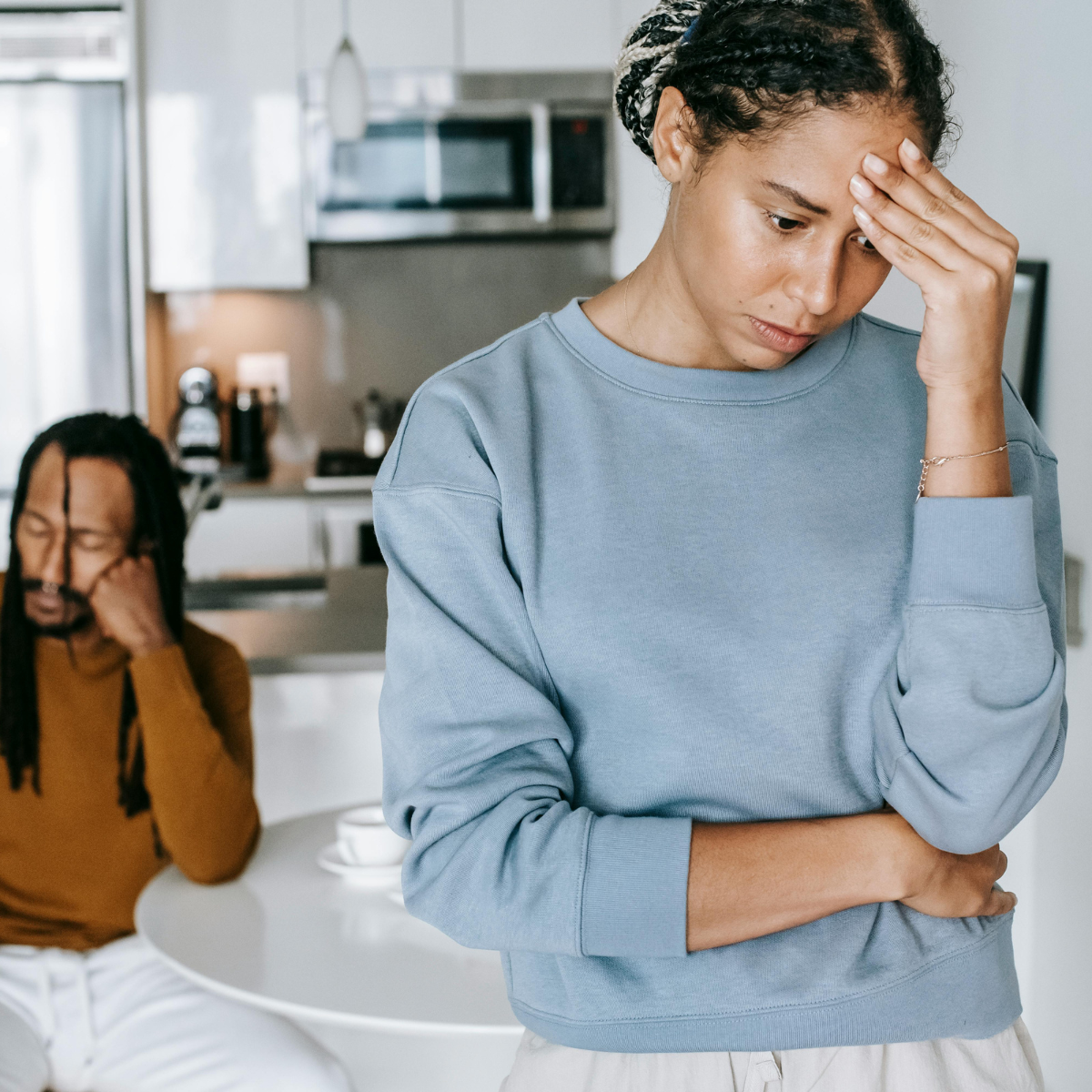 A woman in a blue sweatshirt appears upset, holding her forehead with her hand, while a man with dreadlocks in a brown sweater sits at a table in the background, wearing glasses and appears distressed.