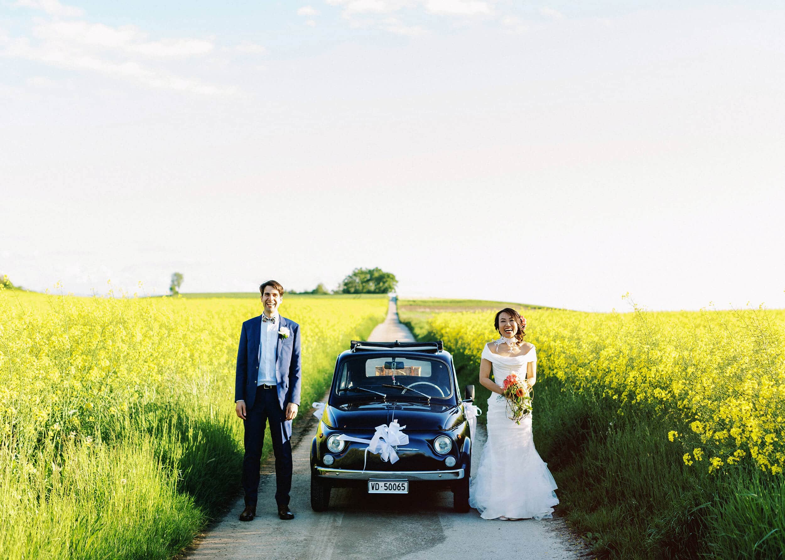 A bride and groom stand beside a vintage car in a bright yellow canola flower field on their wedding day.