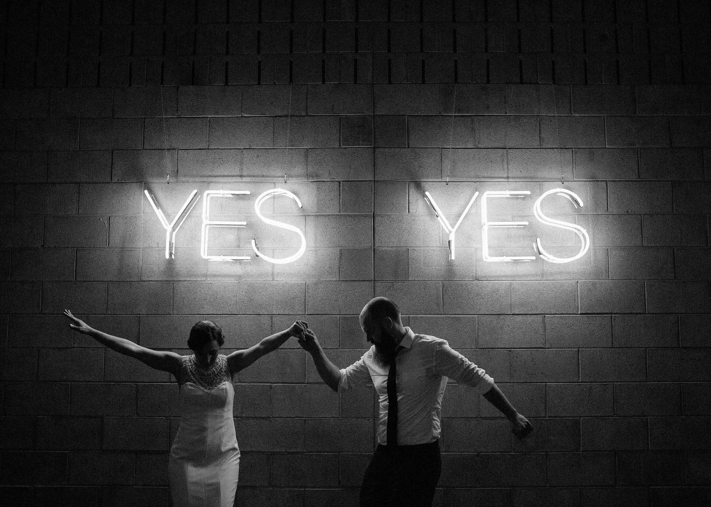 A couple dance beneath glowing neon signs in a candid black-and-white wedding photograph.