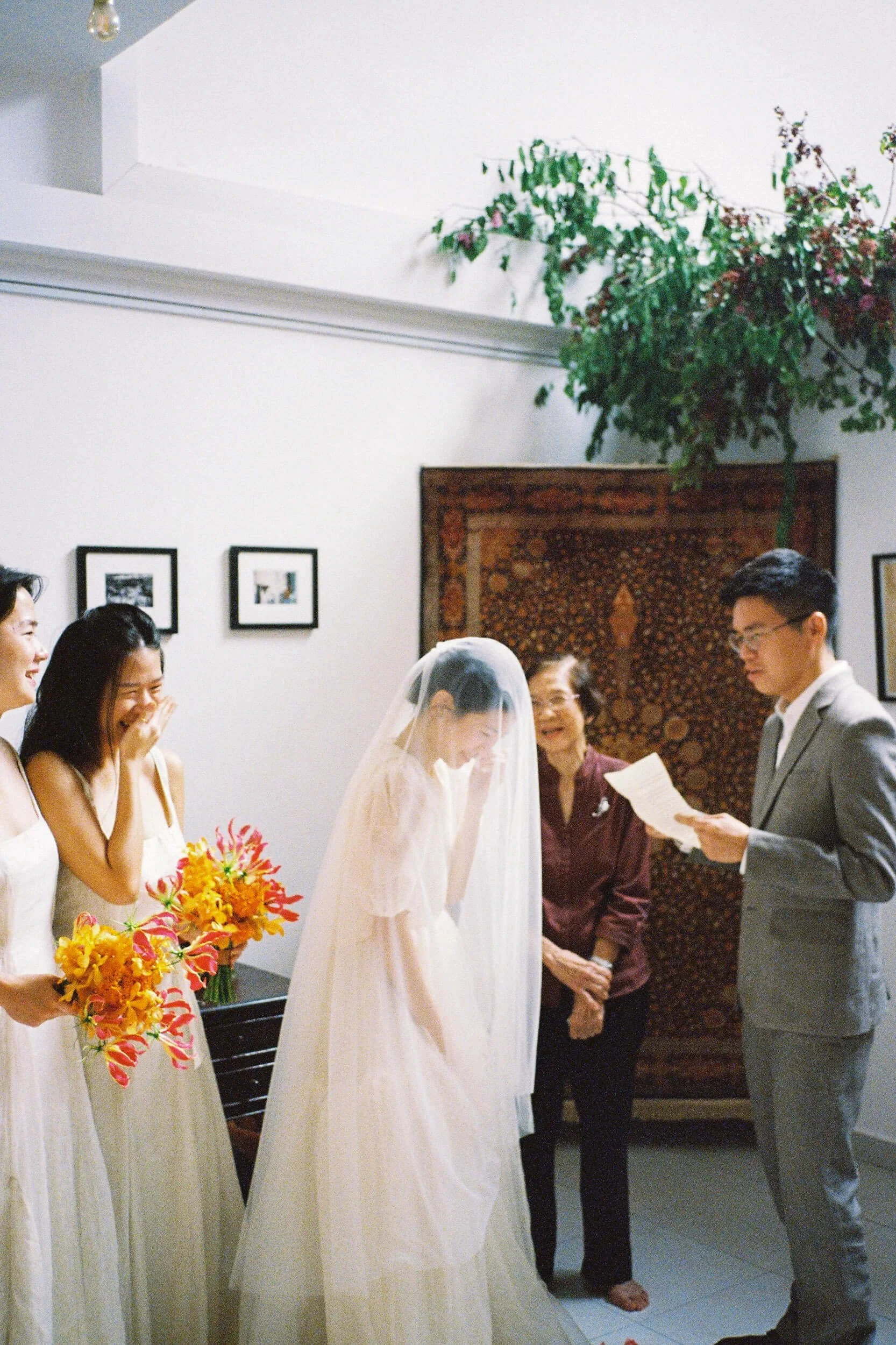 A bride laughs beneath her veil as the groom reads aloud during a warm indoor wedding moment, photographed on 35mm film.