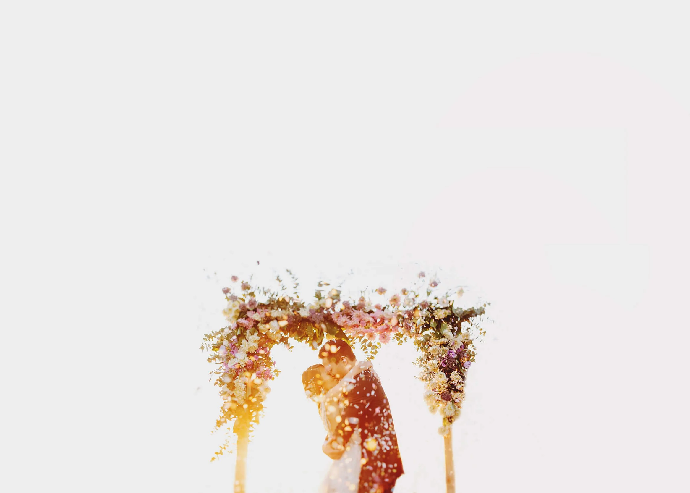 A newlywed couple embrace beneath a floral arch in an airy, whimsical wedding portrait.