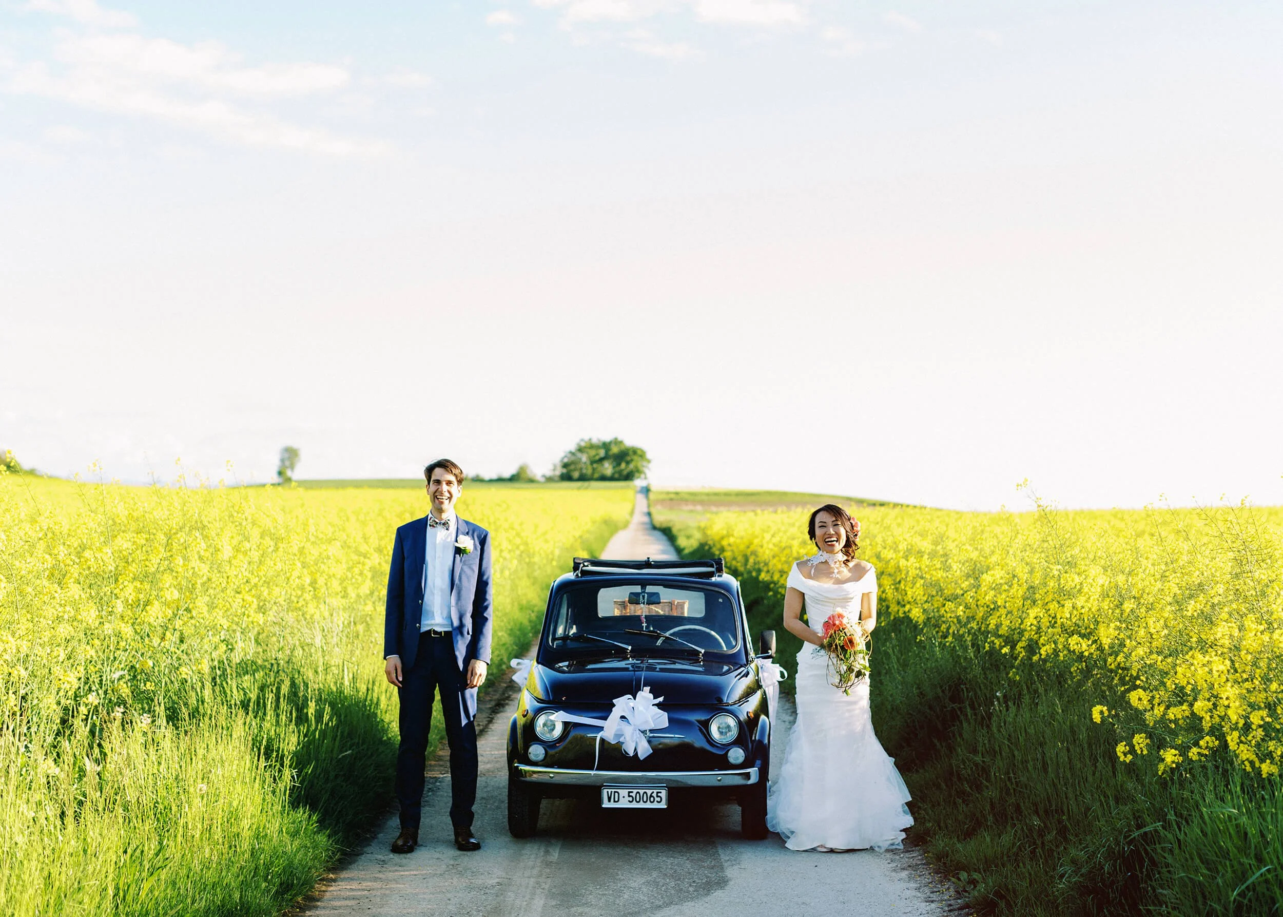 A bride and groom stand beside a vintage car in a bright yellow canola flower field on their wedding day.